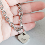 Silver chain bracelet with heart-shaped tag engraved 'Mom' held in a hand.