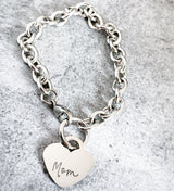 Silver chain bracelet with a heart-shaped tag engraved with 'Mom' on a gray textured background