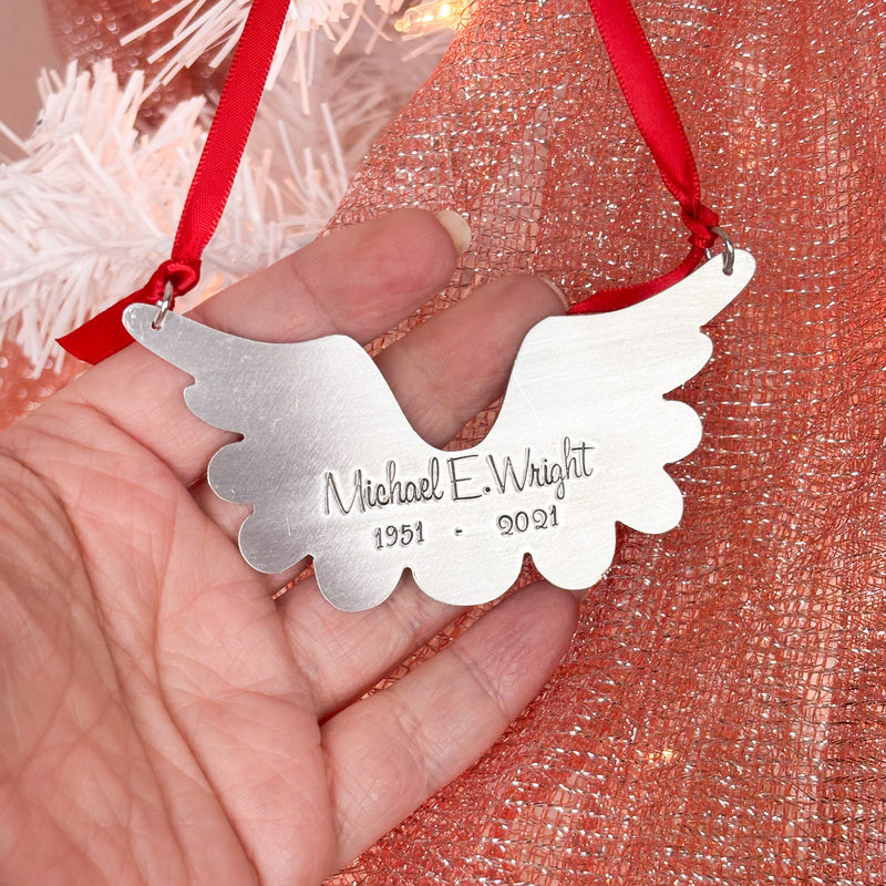 Silver angel wing ornament with engraved text held in a hand against a red and white textured background.