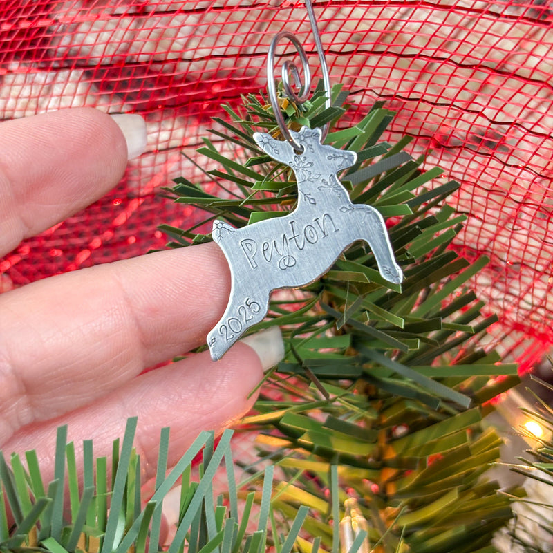 Silver reindeer ornament with name engraved, held over a Christmas tree to show size.